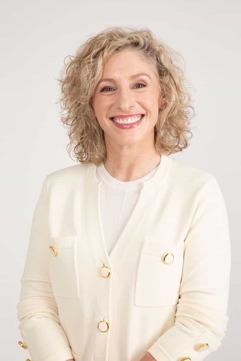 A smiling blonde woman wearing a cream cardigan with gold buttons.