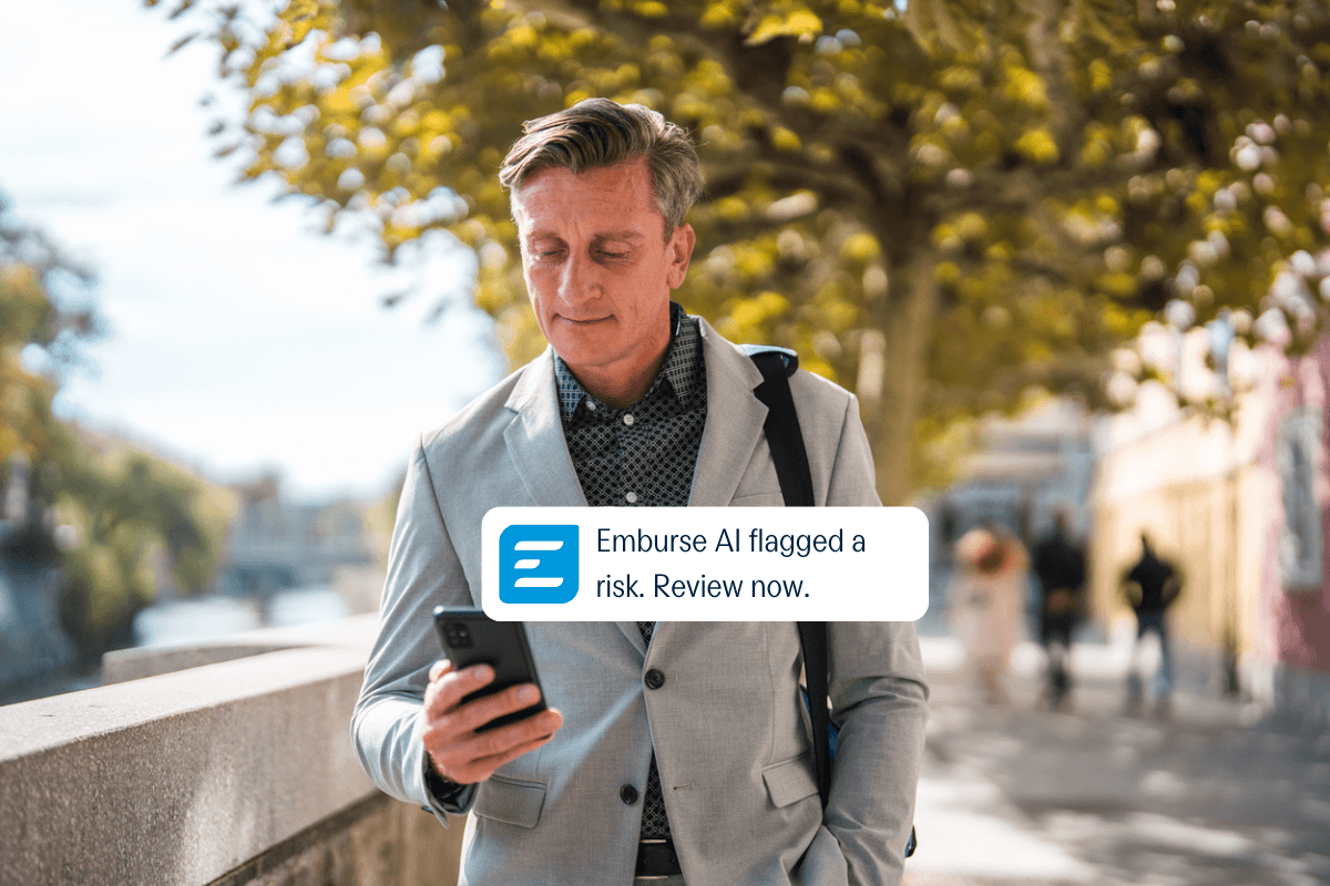 A man outdoors looks at his smartphone showing a notification: "Emburse AI flagged a risk. Review now."